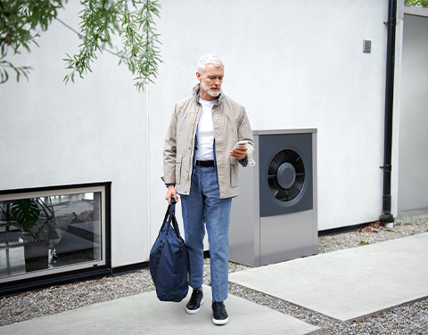 Ein Mann blickt auf sein Smartphone in seiner linken Hand und steht vor einer weißen Hauswand. Im Hintergrund ist eine moderne Wärmepumpe zu erkennen.