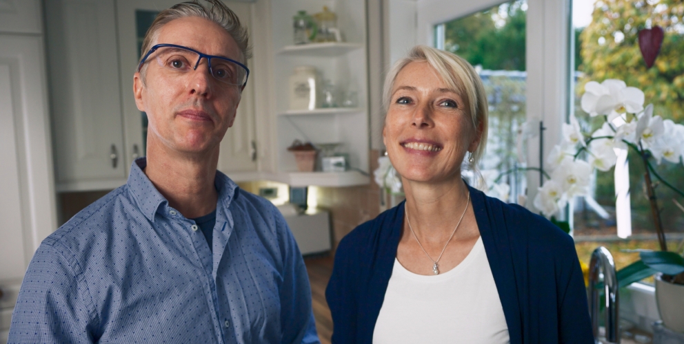 Mann und Frau stehen in einer modernen und hellen Küche. Beide blicken freudig in die Kamera.