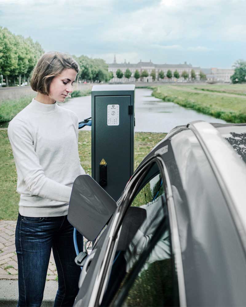 Frau steht an öffentlicher Ladesäule neben ihrem Auto
